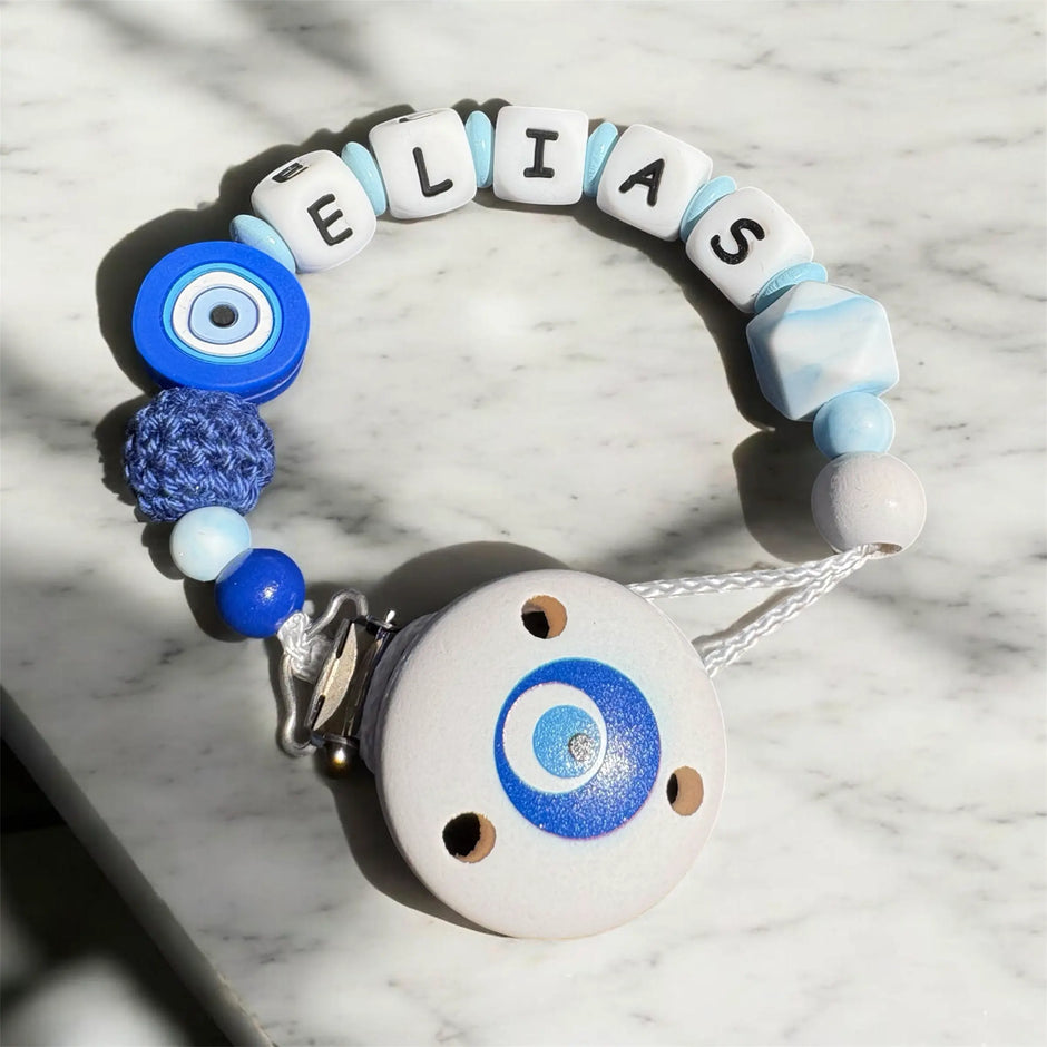 Personalisierte Schnullerkette mit Namen „Elias“ in Blau und Weiß mit Nazar Auge, Holzclip und Silikonperlen auf hellem Marmortisch