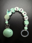 Detailansicht der personalisierten Schnullerkette Dino mit Namen von BabyLions® mit Dino-Motiv, Holzclip, Silikonring sowie Silikon- und Holzperlen in Mint