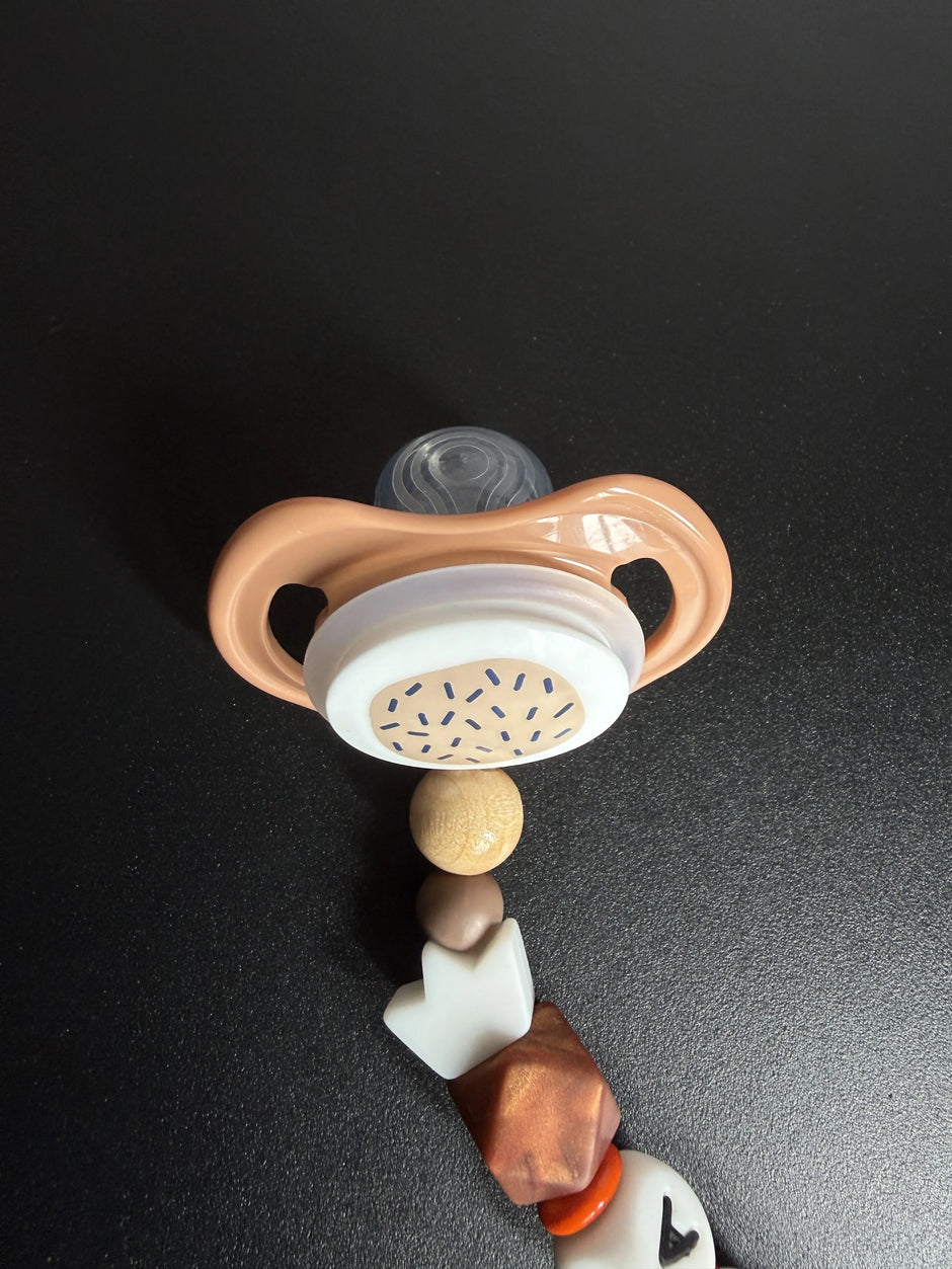 Detailaufnahme eines Babyschnullers an einer personalisierten Schnullerkette in Braun, Beige und Weiß