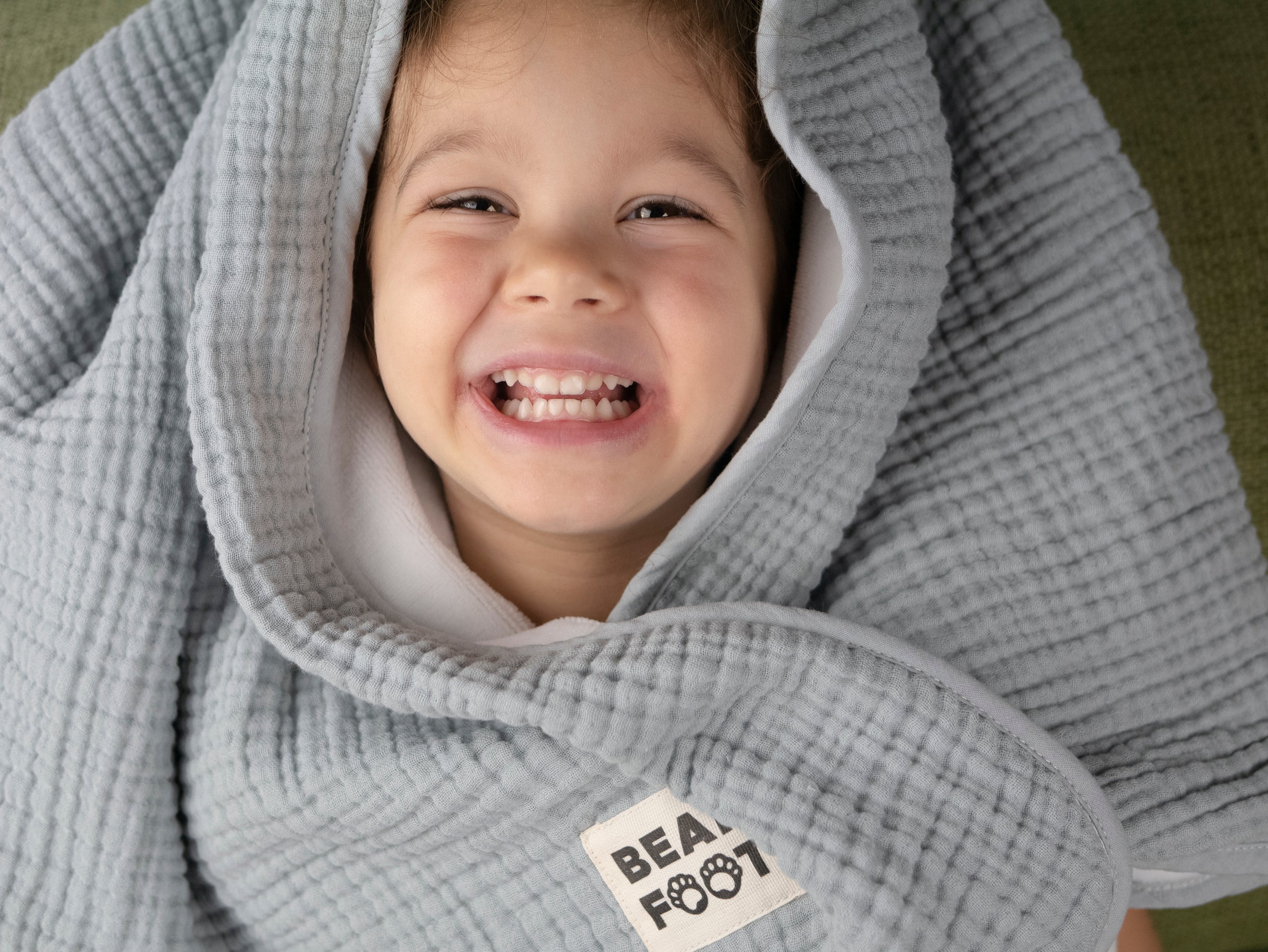 Glückliches Kind eingekuschelt in graue Musselin-Babydecke von Bearfoot – weiche, atmungsaktive Babydecke für Neugeborene und Kleinkinder