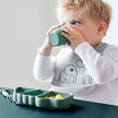 Kleinkind mit weißem Lätzchen mit Elefantenmotiv trinkt aus einem grünen Silikonbecher, vor sich ein grüner Silikon-Stick&Stay-Teller „Crocodile“ mit aufgeschnittenem Obst – erhältlich bei BabyLions.
