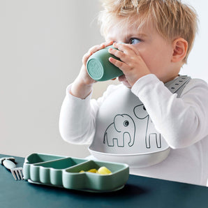 Kleinkind mit weißem Lätzchen mit Elefantenmotiv trinkt aus einem grünen Silikonbecher, vor sich ein grüner Silikon-Stick&Stay-Teller „Crocodile“ mit aufgeschnittenem Obst – erhältlich bei BabyLions.