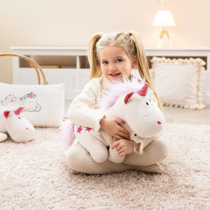 Mädchen mit blonden Zöpfen sitzt auf einem weichen Teppich und umarmt ein weiß-pinkes Kuscheltier-Einhorn „Theodor“ mit rosa Mähne und Sternen – erhältlich bei BabyLions.